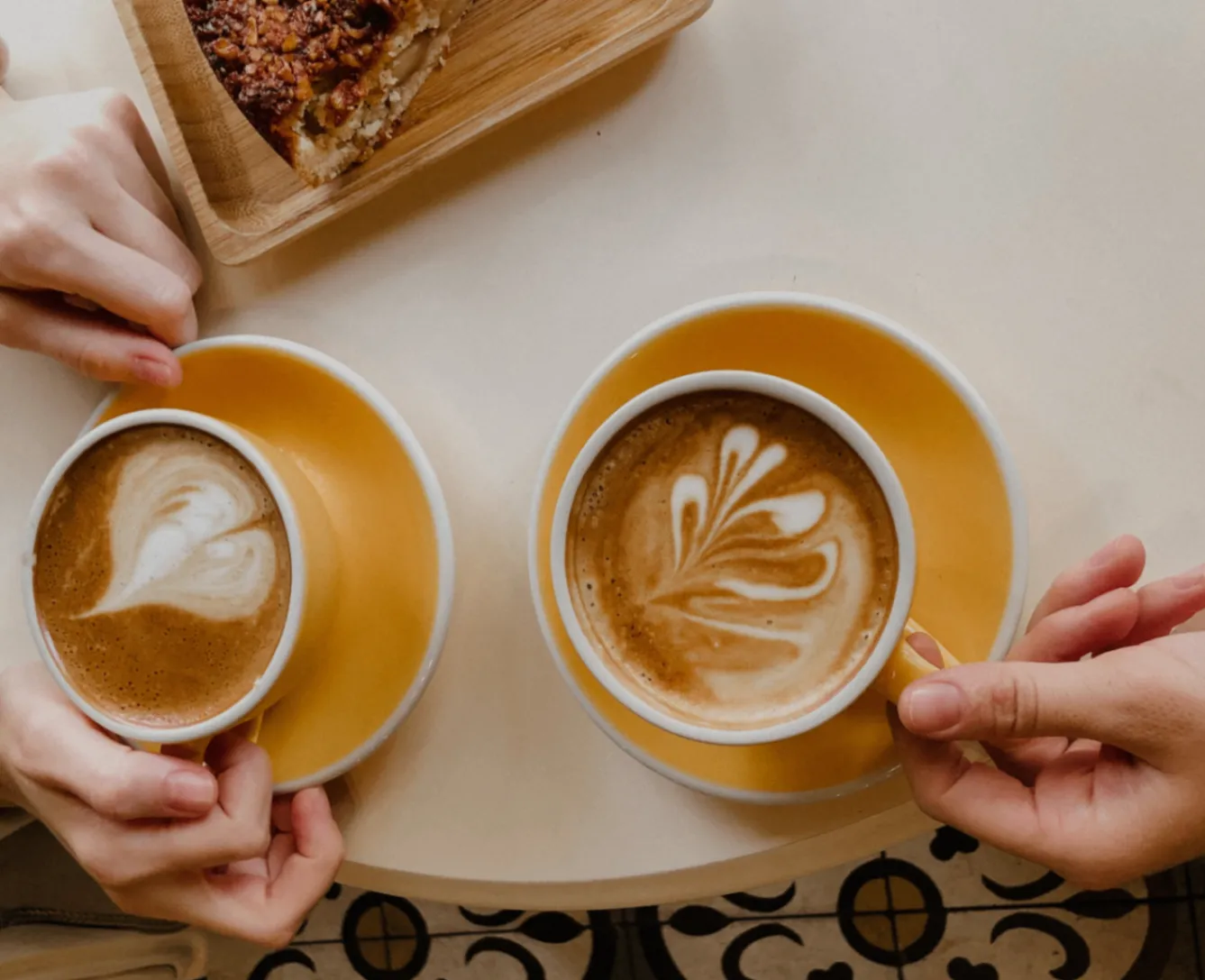 Two hands holding latte art coffee cups over a patterned mosaic table.
